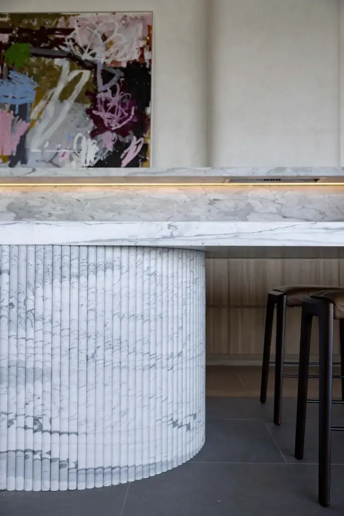 Curved white marble bar with ribbed column base, abstract painting on the wall, and brown stools in a modern interior.