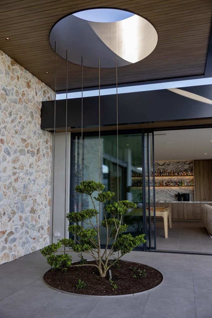 Modern building entrance with a circular skylight in a wooden ceiling, a stone wall, tall hanging metal rods, and open glass doors revealing a bar interior; a circular planter with trimmed shrubs in the foreground.