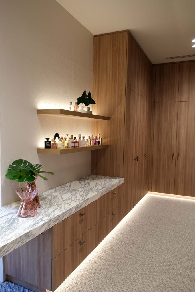 Wood-toned built-in cabinetry with marble countertop, featuring two floating shelves holding perfume bottles and a potted plant under warm lighting.