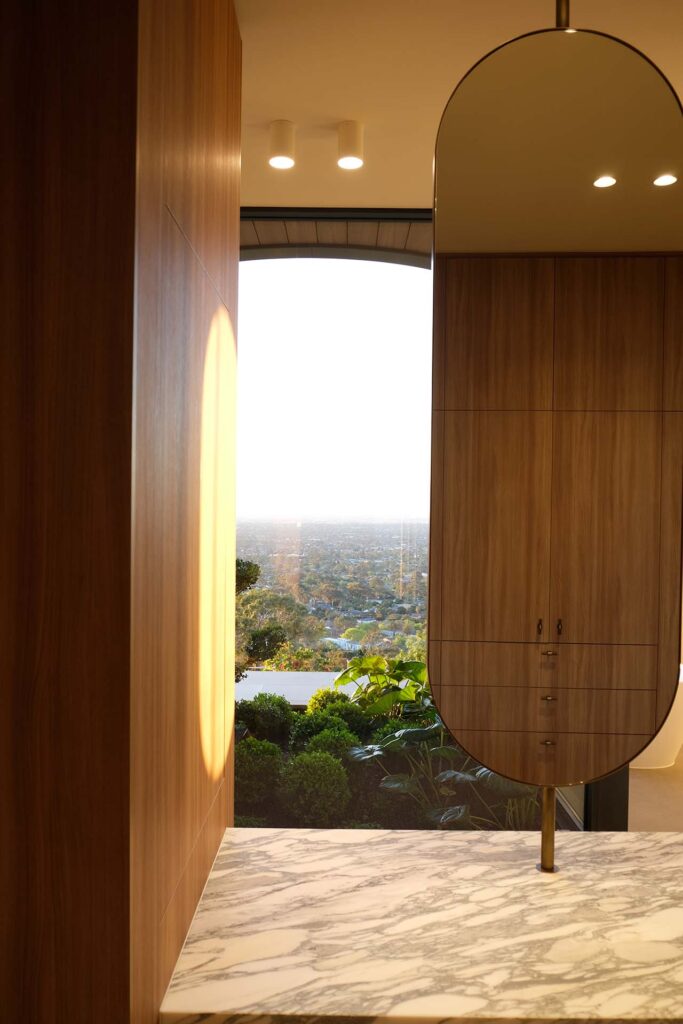 Wood-paneled room with a large oval mirror on the right, marble countertop, and a city view through a window.