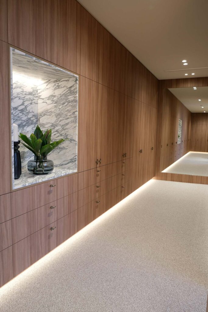 Walnut wood wall cabinets line a hallway with a marble display niche containing a plant and vase, illuminated by built‑in lighting at the base and ceiling spots.