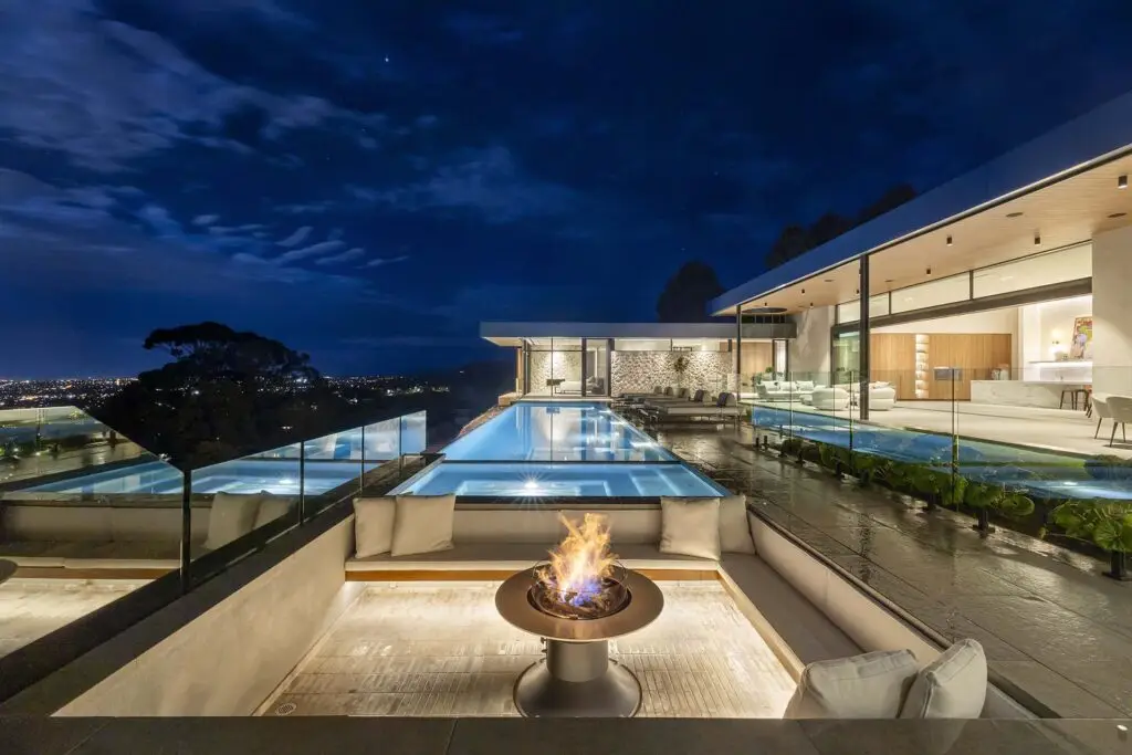 Modern night-time villa with glass-walled living spaces, an illuminated rectangular pool, and a glowing fire pit in a stylish outdoor lounge area overlooking city lights.