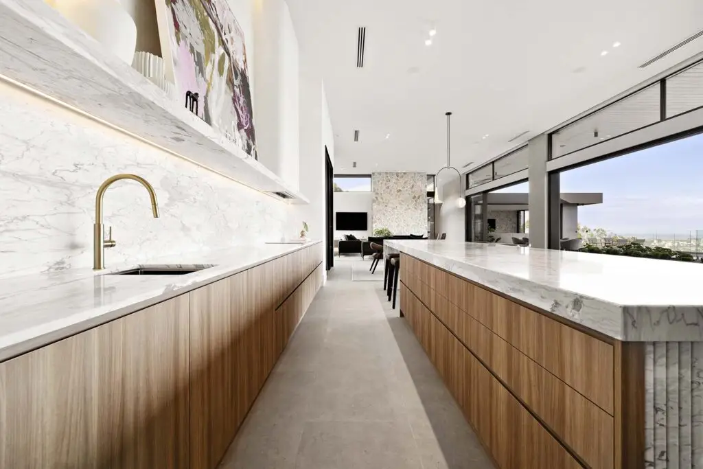 Bright, modern kitchen with marble countertops and a gold faucet, open to a living area and large glass wall overlooking an outdoor scene