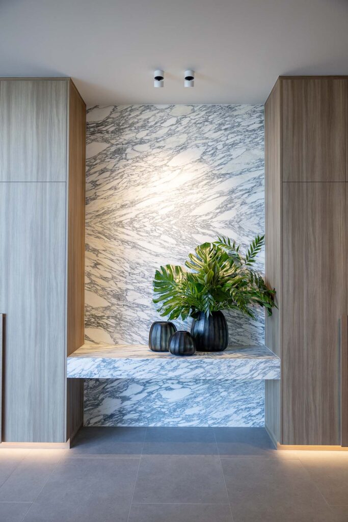 Marble wall niche framed by wood cabinets, with a marble bench holding dark vases and green plants.