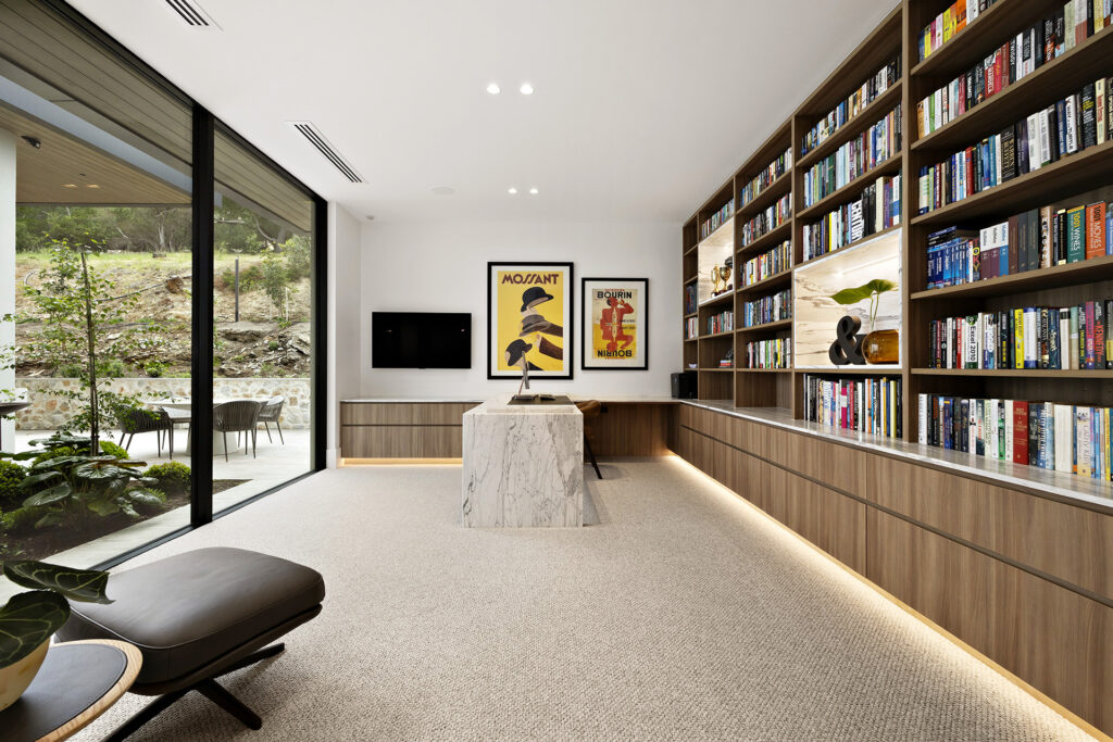 Contemporary home library with tall wooden bookshelves on the right, marble island, and a glass wall opening to an outdoor patio.