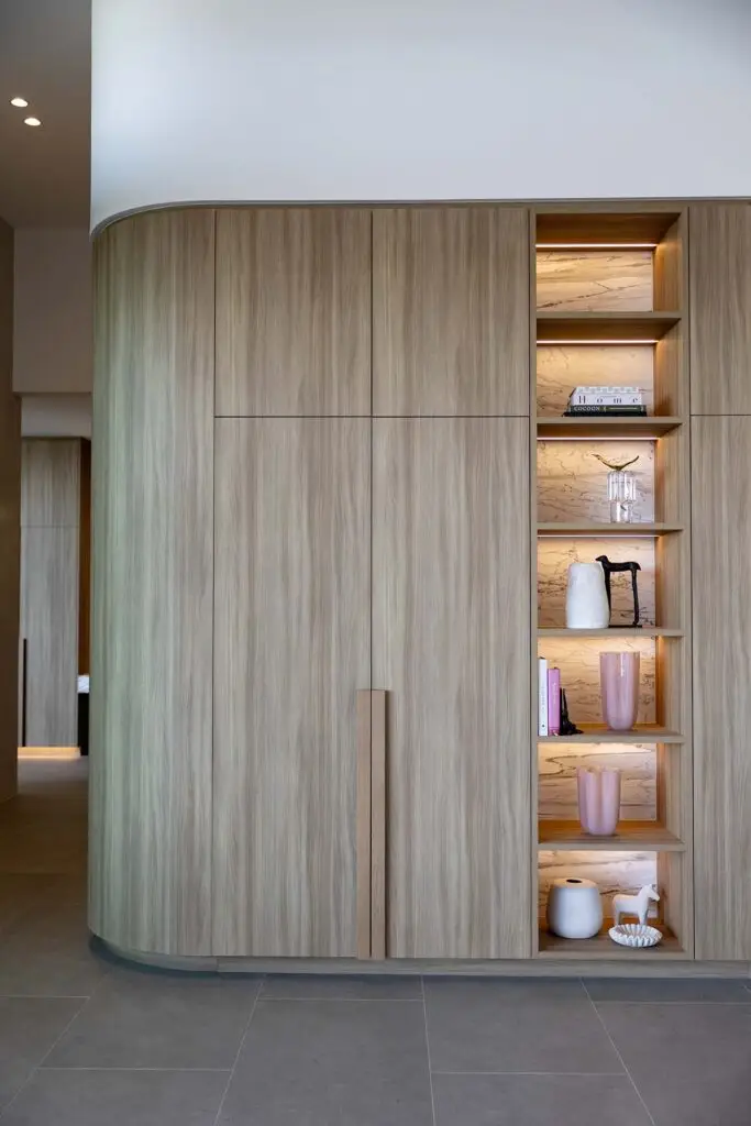 Curved wood cabinet with vertically grained panels and a right-side illuminated display niche showing marble-backed shelves with vases, books, and small sculptures.