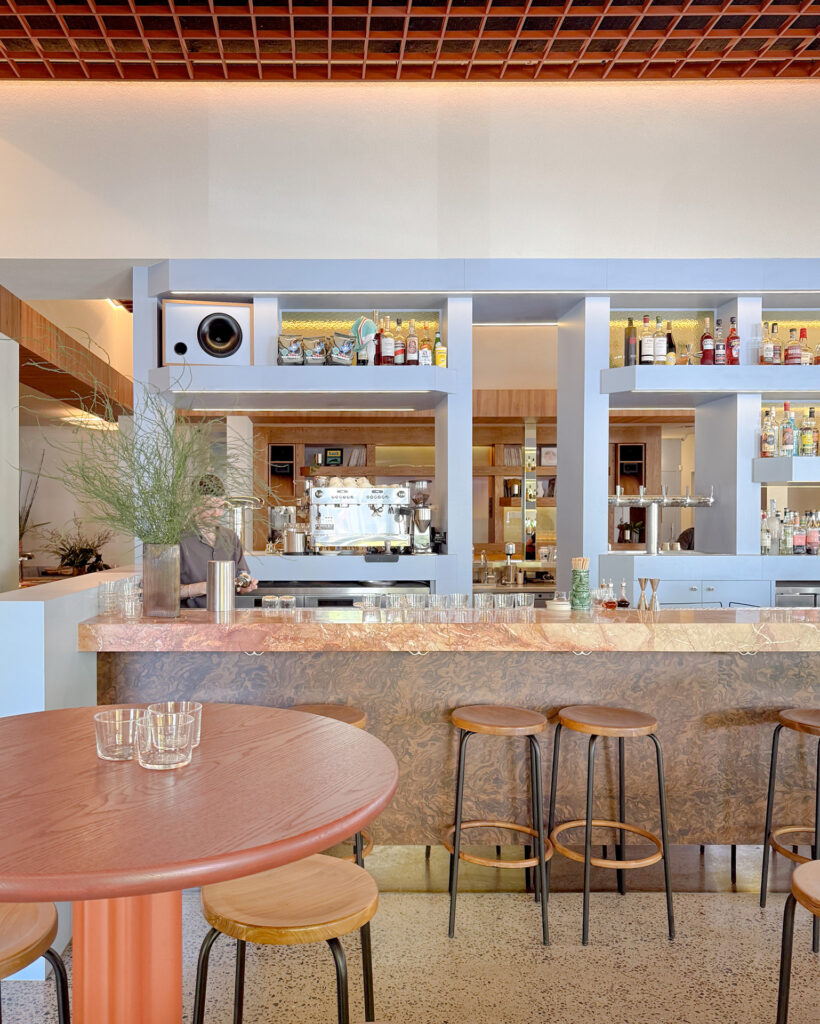 Modern bar interior with a pink marble counter, blue shelving, and bottles on display behind the counter, plus wooden stools in front.