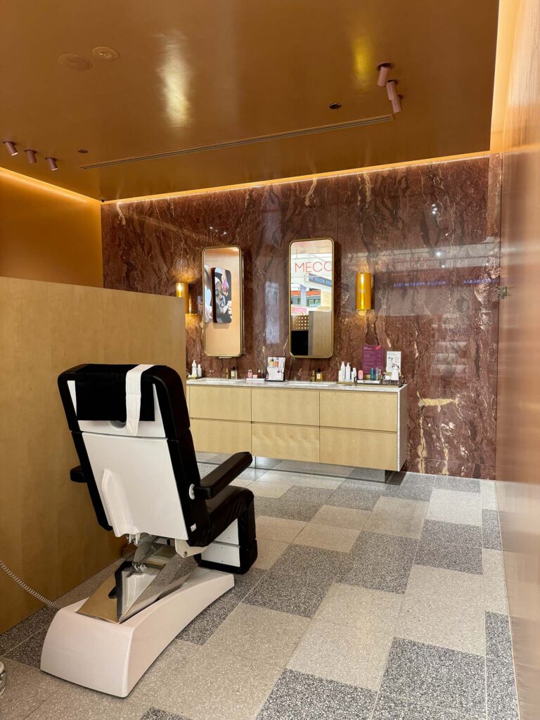 Modern spa/salon interior featuring a white and black reclining chair in front of mirrors and a marble wall with a product-filled vanity visible in the background.