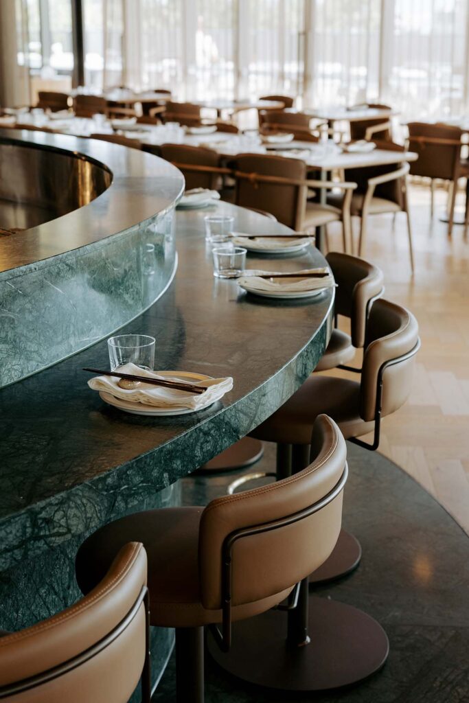 Curved marble bar along a dining area with place settings and glasses at each seat in a sunlit restaurant.