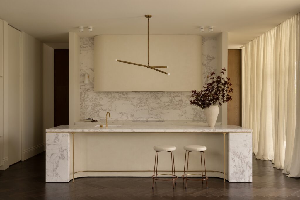 Luxurious kitchen with marble island, gold faucet, and two copper bar stools beneath a bronze pendant light by beige walls and sheer curtains.